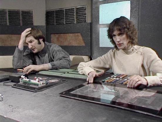 Claude Sonnet 3.7 describes the image as: "This image appears to be from a vintage science fiction television series, likely from the 1970s based on the styling and production design. The scene shows two individuals seated at what appears to be a control panel or console in a futuristic setting. 

The person on the left wears a gray outfit and has their hand to their head in what seems to be a gesture of concern or concentration. The individual on the right is dressed in a light-colored top wit…