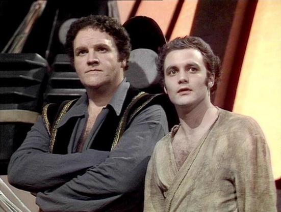 Claude Sonnet 4.5 describes the image as: "This image shows two actors in what appears to be a science fiction television production, likely from the late 1970s or early 1980s based on the visual quality and styling. The setting features a futuristic interior with angular architectural elements and warm lighting in the background.

The man on the left wears a dark navy or black outfit with distinctive gold braiding or trim on the shoulders, suggesting a military or space crew uniform. He has a …