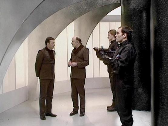 Claude Haiku 4.5 describes the image as: "# Image Description

This scene appears to be from a television production, likely from the 1970s or 1980s based on the styling and image quality. The setting is a futuristic interior space with distinctive curved architectural elements, including arched doorways and a textured dark column on the right side. The walls feature vertical paneling and large windows allowing natural light to flood the space, creating a clean, modernist aesthetic.

Four men a…