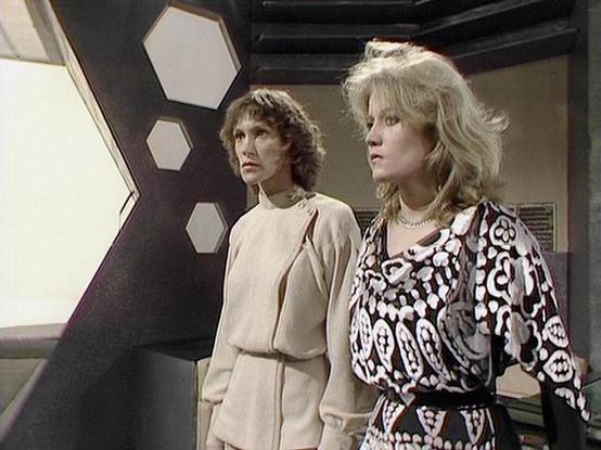 Claude Sonnet 4 describes the image as: "This image shows two women in what appears to be a science fiction television production, likely from the late 1970s or early 1980s based on the styling and video quality. The setting is clearly a futuristic spacecraft or space station interior, characterized by distinctive hexagonal wall panels and sleek, angular design elements typical of sci-fi productions of that era.

The woman on the left wears a simple, elegant cream-colored outfit with clean line…