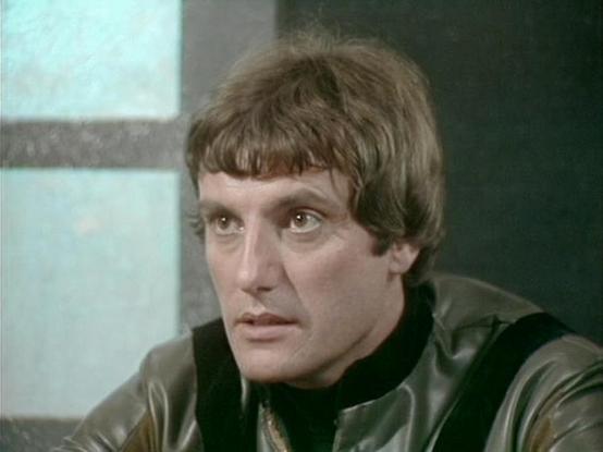 Claude Sonnet 4 describes the image as: "This image shows a scene from what appears to be a science fiction television production, likely from the late 1970s or early 1980s based on the visual style and production quality. The setting appears to be inside a futuristic spacecraft or space station, with characteristic metallic walls and geometric window panels visible in the background.

The person in the image is wearing a distinctive black leather outfit with metallic studs or decorative elemen…