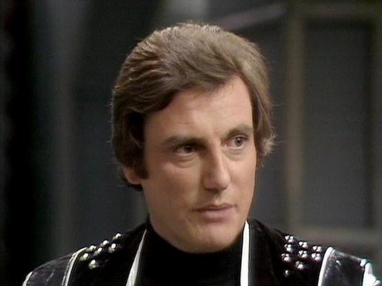 Claude Sonnet 3.7 describes the image as: "The image shows a person with brown hair wearing a distinctive black outfit with studded shoulder details. They appear to be in a scene from what looks like a television production, likely from the late 1970s or early 1980s based on the styling and video quality. The person is wearing a black turtleneck under what appears to be a leather or vinyl costume with metallic studs or embellishments.

The setting has a dark, somewhat industrial background that…