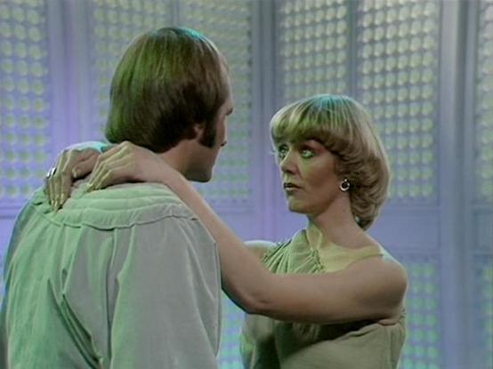 Claude Sonnet 4 describes the image as: "This appears to be a scene from a science fiction television production, likely from the late 1970s or early 1980s based on the styling and video quality. The setting features a futuristic interior with geometric patterned walls or screens in the background, creating a distinctly sci-fi atmosphere with cool lighting tones.

Two characters are shown in an intimate moment, with their arms around each other in what appears to be an emotional or romantic sce…