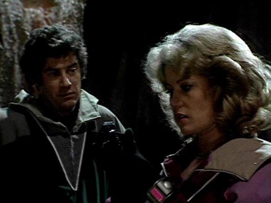 Claude Sonnet 4 describes the image as: "This image appears to be from a science fiction television production, likely from the late 1970s or early 1980s based on the visual style and production quality. The scene shows two characters in what appears to be a dark, industrial or underground setting with rough stone or concrete walls visible in the background.

The male character on the left wears dark, practical clothing with what appears to be some kind of communication device or equipment. The…