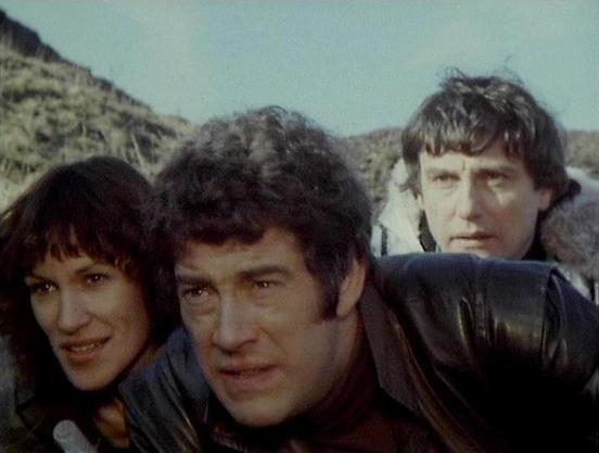 Claude Sonnet 4 describes the image as: "This image appears to be from the British science fiction television series "Blake's 7," which aired from 1978 to 1981. The scene shows three characters in what appears to be an outdoor, rocky terrain setting with hills or mountains visible in the background. The lighting and film quality suggest this is from the late 1970s/early 1980s television production era.

The characters are wearing practical, somewhat utilitarian clothing typical of the show's ae…