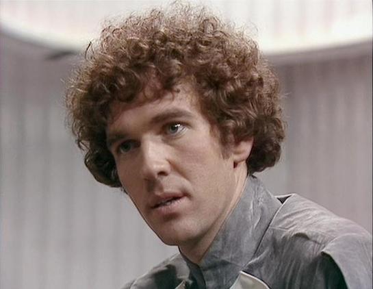 Claude Sonnet 4 describes the image as: "I can see this is an image of a person with curly brown hair wearing what appears to be a light-colored shirt or jacket. The setting looks like it could be from a television production or film, with soft lighting and what seems to be an indoor environment in the background. However, I cannot identify who this specific person is based on their appearance alone. If you'd like to discuss details about Steven Pacey's work or career, I'd be happy to help with…