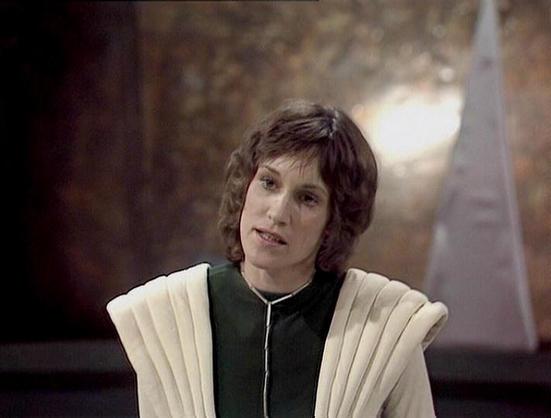 Claude Sonnet 3.7 describes the image as: "The image shows a person with short, dark curly hair wearing a distinctive costume that appears to be from a science fiction production. They're dressed in a dark green or black outfit with a striking cream-colored pleated collar or shoulder piece that fans out dramatically. The background is softly blurred, suggesting an indoor set with subtle lighting.

This appears to be a screenshot from what looks like a classic science fiction television producti…