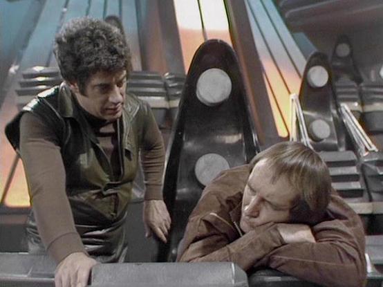 Claude Sonnet 4 describes the image as: "This image shows a scene from the classic British science fiction series "Blake's 7" (1978-1981). The setting appears to be the interior of a spacecraft, likely the Liberator, with its characteristic futuristic control room featuring curved walls, metallic surfaces, and distinctive lighting. The scene shows two characters in what appears to be a tense or concerning moment - one person leaning over another who appears to be unconscious or resting on what …