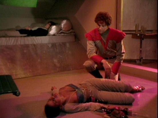 Claude Haiku 4.5 describes the image as: "# Scene Description

This appears to be a dramatic scene set in a futuristic spacecraft interior, characterized by the distinctive production design typical of 1980s science fiction television. The setting features angular walls, metallic surfaces, and distinctive colored lighting that creates an otherworldly atmosphere.

The scene depicts an intense moment of conflict or crisis. One character lies on the floor in a vulnerable position, while another ch…