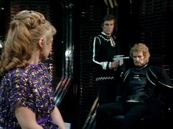 Claude Sonnet 4 describes the image as: "This appears to be a scene from a science fiction television production, likely from the late 1970s or 1980s based on the visual style and production quality. The setting appears to be the interior of a spacecraft or futuristic facility, with dark metallic walls and technical equipment visible in the background.

The scene shows three people in what appears to be a tense dramatic moment. On the left is a woman with an elaborate blonde updo hairstyle, wea…