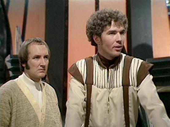 Claude Sonnet 4.5 describes the image as: "This image appears to be from a television production, likely from the late 1970s or early 1980s based on the video quality and styling. Two men are shown in period costume suggesting a historical or science fiction setting. 

The actor on the left wears a simple cream-colored tunic with a beige vest or overdress, giving a rustic, peasant-like appearance. His hair is styled in a practical, short cut appropriate to the period costume.

The actor on the …