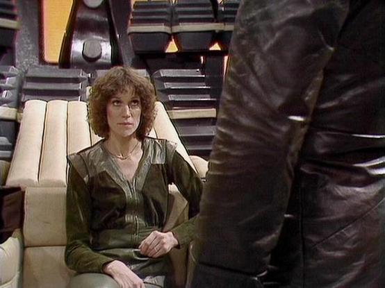 Claude Haiku 4.5 describes the image as: "# Image Description

This scene appears to be from a science fiction television production, set within a spaceship interior. The setting features a sleek, futuristic design with cream-colored seating, dark control panels, and ambient lighting typical of 1970s-80s sci-fi aesthetics. The architectural elements include geometric shapes and a modernist bridge or upper structure visible in the background.

In the foreground, a woman with curly blonde hair si…