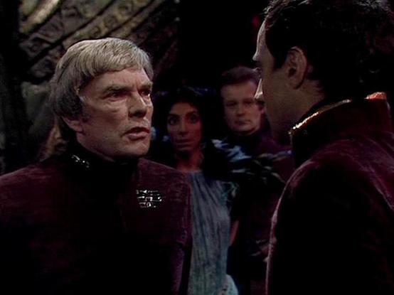 Claude Sonnet 4.5 describes the image as: "This image appears to be from a classic science fiction television production, likely from the 1970s or 1980s based on the video quality and costume design. The scene is dimly lit with dramatic lighting that creates a tense, confrontational atmosphere.

The setting appears to be an ornate interior space with decorative elements visible in the background, suggesting a formal or ceremonial location. The characters are wearing elaborate burgundy and dark-…