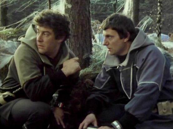 Claude Sonnet 4 describes the image as: "This image shows two men in a forest setting, sitting among fallen logs and woodland debris. They're wearing practical outdoor clothing - one in an olive/brown hooded jacket and the other in a blue/grey hoodie. The setting appears to be from a film or television production, with the natural forest environment providing a rustic backdrop. The lighting and image quality suggest this is from an older production, likely from the 1970s or 1980s. The scene has…