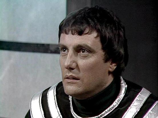 Claude Haiku 4.5 describes the image as: "# Image Description

This image shows an actor in character wearing a distinctive black and white striped tunic or uniform with metallic silver trim and accents around the neckline and shoulders. The costume suggests a science fiction setting, likely from a television production. The character has dark hair styled in a distinctive 1970s cut with bangs, and displays a serious, somewhat intense expression.

The setting appears to be an interior space with…