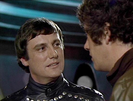 Claude Haiku 4.5 describes the image as: "# Scene Description

This image appears to be from a science fiction television production, showing two men in conversation. The figure on the left wears a distinctive black leather jacket adorned with metal studs and rivets, which appears to be a futuristic costume design. The lighting is dramatic, with blue-tinted atmospheric lighting visible in the background, suggesting an interior spacecraft or sci-fi setting.

The scene captures an intimate moment…