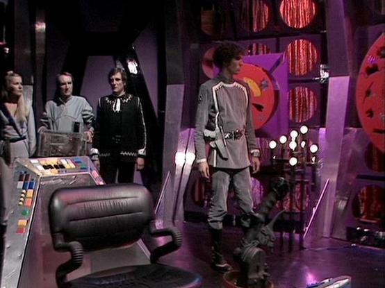 Claude Sonnet 4 describes the image as: "This appears to be a scene from the British science fiction television series "Blake's 7," showing the characteristic futuristic set design with purple and pink lighting that was typical of the show's aesthetic. The scene takes place on what appears to be the flight deck or control room of a spacecraft, with the distinctive curved control console and technological equipment visible in the foreground. Several crew members are positioned around the set in …