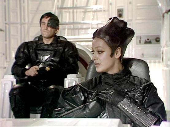 Claude Sonnet 4 describes the image as: "This image appears to be from a science fiction television production, likely from the 1970s or 1980s based on the visual style and production values. The scene takes place in what looks like a futuristic spacecraft or space station interior, with clean white walls and modern-looking furniture/seating.

Two figures are visible wearing distinctive black leather or vinyl uniforms with military-style details and metallic accents. The costumes have a sleek, …