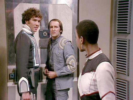 Claude Haiku 4.5 describes the image as: "# Scene Description

This image captures a tense moment in what appears to be a spacecraft corridor or futuristic setting. Three characters are engaged in conversation, standing in a metallic, industrial-style environment with distinctive architectural features including ventilation grilles and paneled walls typical of science fiction production design.

The scene conveys dramatic tension, with the characters positioned in a triangular formation that su…