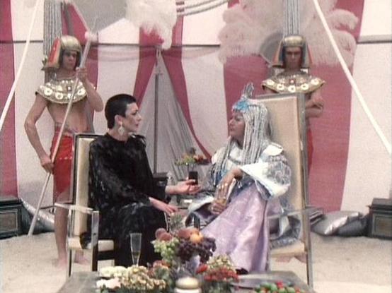 Claude Haiku 4.5 describes the image as: "# Egyptian-Themed Scene Description

This image shows a theatrical scene set in ancient Egypt, featuring elaborate production design with distinctive Egyptian iconography and styling. The setting includes ornate backdrops with hieroglyphic-inspired patterns and Egyptian motifs in shades of pink, red, and gold.

Two central figures are seated in conversation, dressed in period costume. One wears a black outfit while the other is adorned in an elaborate E…