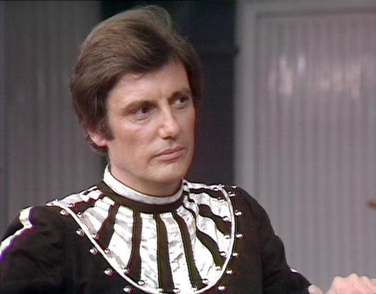 Claude Sonnet 4.5 describes the image as: "This image shows a dramatic scene from what appears to be a British science fiction television production, likely from the late 1970s or early 1980s. The actor is wearing an elaborate costume featuring a striking black garment with a distinctive white and brown geometric collar piece that creates a bold, futuristic aesthetic. The collar has a segmented, almost armor-like quality with metallic studs or rivets adorning it.

The setting appears to be an i…