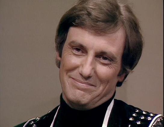 Claude Sonnet 3.7 describes the image as: "This image shows a person with brown hair styled in a 1970s-80s fashion, wearing what appears to be a dark outfit with some decorative elements or embellishments visible at the shoulder. They have a distinctive slight smirk or knowing smile on their face against a neutral background. 

The styling, costume, and filming quality suggest this is from a television production from that era. The dark costume with decorative elements could indicate this is fr…