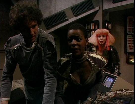 Claude Sonnet 4 describes the image as: "This appears to be a scene from a science fiction television production, likely from the 1980s based on the visual style and production values. The setting is clearly a futuristic spacecraft or space station interior, with characteristic sci-fi elements like computer terminals, metallic surfaces, and technical equipment visible in the background.

The scene shows three characters in what appears to be a tense or dramatic moment. One figure is wearing a l…