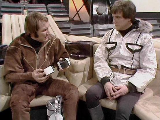 Claude Sonnet 4.5 describes the image as: "This image shows a scene from the British science fiction television series "Blake's 7." Two actors are seated on a beige curved sofa in what appears to be a spaceship interior set. The setting features a futuristic design with metallic elements and technical equipment visible in the background.

On the left, one actor wears a brown leather-like jacket and is holding what appears to be small cards or devices in his hands. On the right, the other actor …