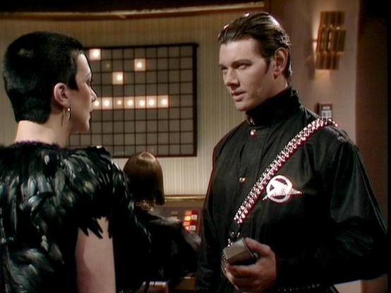 Claude Sonnet 4 describes the image as: "This image shows a scene from what appears to be a science fiction production, likely from the 1970s or 1980s based on the aesthetic and production values. The setting appears to be an interior space with warm lighting and geometric design elements visible in the background, including what looks like a grid pattern with illuminated squares.

Two figures are visible in the scene, both wearing distinctive dark costumes that suggest this is from a dramatic …