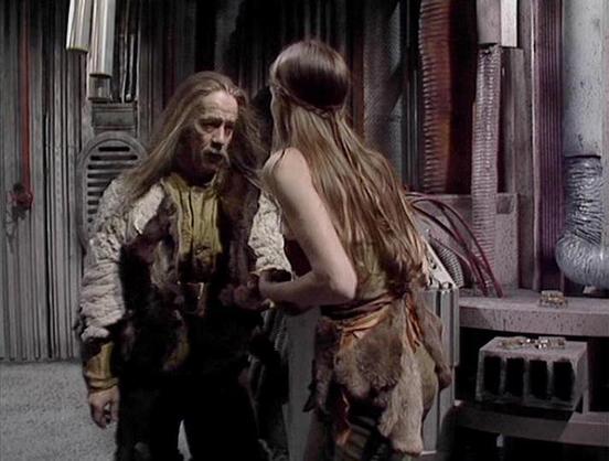 Claude Sonnet 3.7 describes the image as: "This image appears to be from a science fiction or fantasy television production, likely from the late 1970s or early 1980s based on the visual style. The scene takes place in what looks like an industrial or spacecraft interior setting with metallic walls and pipes visible in the background.

Two figures are shown in conversation. They're wearing primitive-looking costumes made of fur and rough fabric, which creates an interesting contrast with the te…