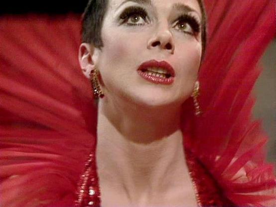 Claude Sonnet 4 describes the image as: "I can see this is a dramatic theatrical or film scene featuring an elegant performer in what appears to be a striking red costume or outfit with feathered or fan-like decorative elements creating a dramatic backdrop. The person is wearing bold red lipstick and ornate jewelry, with their hair styled in a sleek, vintage-inspired look. The lighting and composition create a very theatrical, possibly cabaret or performance art atmosphere. The dramatic makeup,…