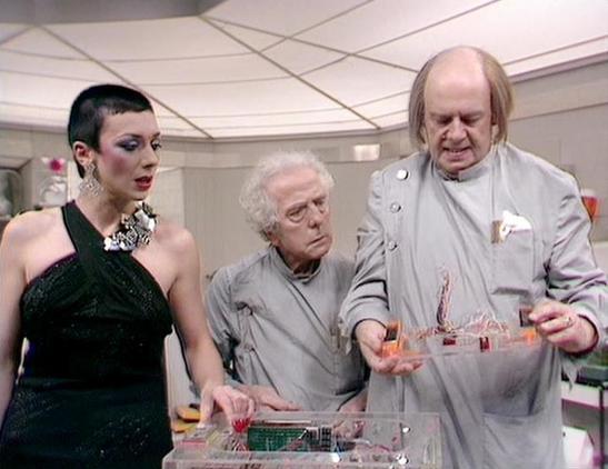 Claude Sonnet 4.5 describes the image as: "This appears to be a behind-the-scenes or production still from what looks like a science fiction television series, likely from the late 1970s or early 1980s based on the aesthetic and set design. The scene is set in what appears to be a futuristic control room or spaceship interior, characterized by white walls, a geometric ceiling panel design, and technical equipment visible in the foreground.

Three actors are positioned around what seems to be a …