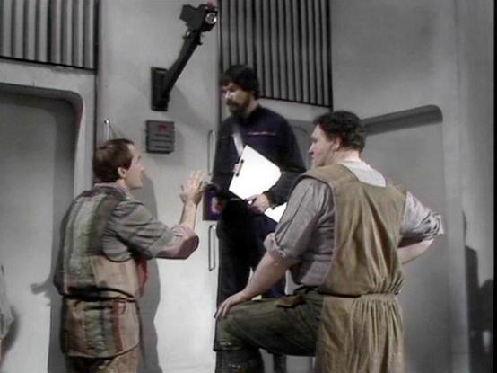 Claude Sonnet 4.5 describes the image as: "This image appears to be from the British science fiction television series "Blake's 7" (1978-1981). The scene shows three characters in what appears to be a corridor or interior space of a spacecraft, characterized by typical BBC sci-fi set design of that era with grey walls and industrial-looking panels.

Two actors are visible in period-appropriate costumes consisting of earth-toned tunics and practical garments typical of the show's aesthetic. One …