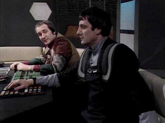 Claude Sonnet 4.5 describes the image as: "This image shows a scene from the British science fiction television series "Blake's 7," which aired from 1978 to 1981. The setting appears to be the flight deck of the spacecraft Liberator, characterized by its distinctive retro-futuristic design with dark walls, geometric light panels, and console stations featuring colorful buttons and controls.

Two crew members are seated at the control stations. On the left is Michael Keating, wearing a brown and…