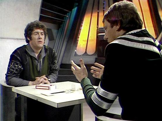 Claude Sonnet 3.7 describes the image as: "This image shows a scene from what appears to be a classic science fiction television production, likely from the British series "Blake's 7." The setting features a futuristic interior with distinctive colored panels visible in the background.

Two people are engaged in conversation across a white table or console. The person on the left wears a dark sweater with patterned sleeves, while the person on the right is dressed in a striped outfit with disti…