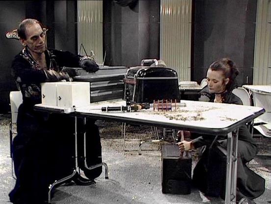 Claude Sonnet 4.5 describes the image as: "This image shows a scene from what appears to be a science fiction television production, likely from the 1970s based on the aesthetic and production design. Two actors are seated at opposite ends of a white table in what looks like a futuristic or institutional setting with dark walls and metallic paneling.

The setting features modernist furniture including black office chairs and a white work surface. On the table are various props including what ap…