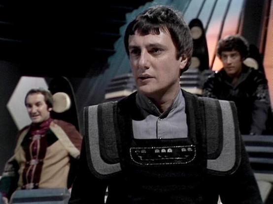 Claude Sonnet 3.7 describes the image as: "This image appears to be from the classic British science fiction television series "Blake's 7." The scene is set in what looks like a spaceship control room or bridge, with futuristic design elements typical of the show's distinctive visual style. 

The image features performers in costume wearing the characteristic styling of the series. The person in the foreground is wearing a distinctive outfit with padded shoulders and a gray and black uniform wi…