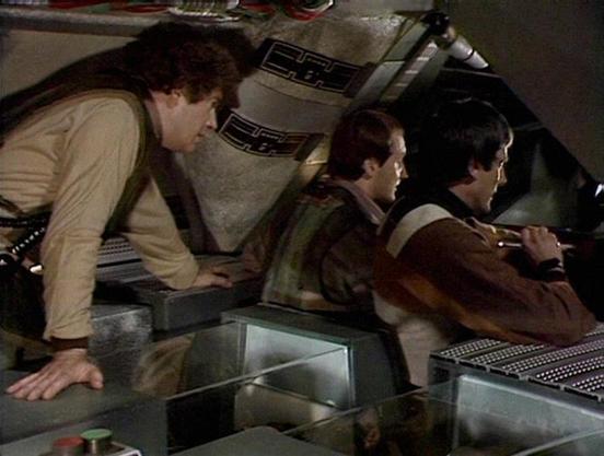 Claude Haiku 4.5 describes the image as: "# Cockpit Scene from Blakes 7

This image shows three actors in the cramped cockpit of a spacecraft, depicting a tense moment during the classic British science fiction series. The setting is the interior of a starship, with control panels and technical equipment visible in the background. The men are dressed in utilitarian flight suits appropriate to the show's aesthetic.

The scene captures the characteristic low-budget production design of 1970s Brit…