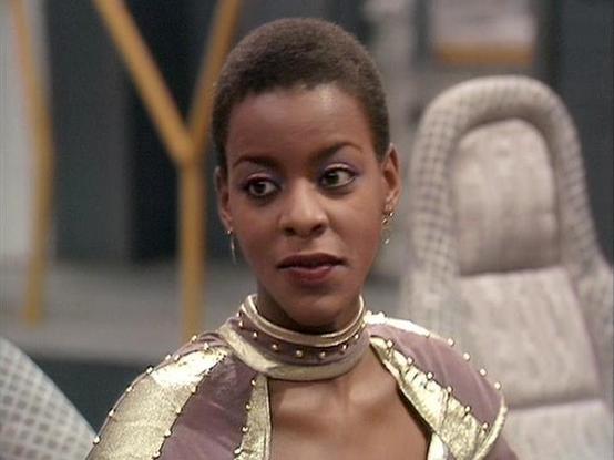 Claude Sonnet 4.5 describes the image as: "This image shows a scene from what appears to be a science fiction television production, likely from the 1980s based on the visual style and production quality. The actor Josette Simon is featured in an elaborate costume that suggests a regal or warrior character. She wears ornate golden armor with studded brown leather elements and multiple gold neck rings that create a striking collar effect. Her makeup includes dramatic eye definition, and she has …