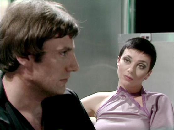 Claude Haiku 4.5 describes the image as: "# Scene Description

This image captures a dramatic moment between two characters in what appears to be an interior setting with institutional or clinical aesthetics, suggested by the plain walls and modern furnishings visible in the background. The scene has the characteristic look of 1970s television production quality.

On the left, one character is shown in profile, wearing a dark garment with shoulder-length dark hair styled in the fashion typical …