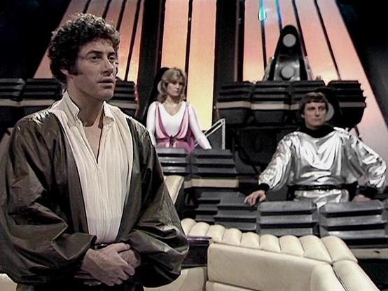 Claude Haiku 4.5 describes the image as: "# Blakes 7 Scene

This image captures a scene from the British science fiction series "Blakes 7," set within a futuristic spacecraft interior. The set design features striking angular architecture with curved metallic panels and a distinctive color-graded backdrop of warm peachy-orange tones that create an otherworldly atmosphere.

In the foreground, an actor portrays a character in a simple brown tunic-style outfit, standing with a contemplative expres…