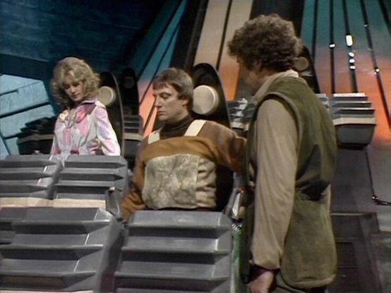 Claude Sonnet 4 describes the image as: "This image appears to be from the British science fiction television series "Blake's 7" from the late 1970s/early 1980s. The scene is set in what looks like a futuristic spacecraft interior with metallic steps and control panels. The setting has the characteristic low-budget but imaginative production design typical of British sci-fi from that era.

The scene shows three characters in a tense moment on what appears to be the flight deck or control area o…