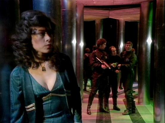 Claude Haiku 4.5 describes the image as: "# Blake's 7 Scene Description

This image appears to be from the science fiction television series "Blake's 7," showcasing a dramatic interior spaceship setting. The scene features characters in a futuristic corridor with distinctive red-toned architectural elements and green-tinted lighting effects that create an otherworldly atmosphere.

In the foreground, a woman with dark, voluminous hair wears a striking teal-blue tunic with ornate gold trim and a …