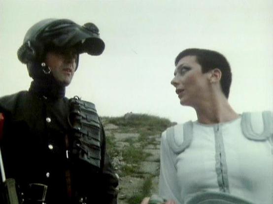 Claude Sonnet 4 describes the image as: "This image appears to be from a science fiction television production, likely from the 1970s or 1980s based on the visual style and film quality. The scene shows two characters in what appears to be an outdoor, rocky or mountainous setting. One figure is dressed in dark, military-style clothing with ornate details and a distinctive cap or headpiece, while the other wears a light-colored, futuristic-looking garment with metallic or reflective elements. Th…