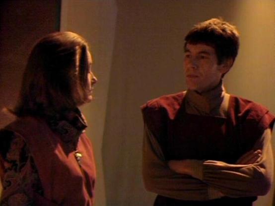 Claude Sonnet 3.7 describes the image as: "The image shows a scene from what appears to be a science fiction production, with two people in a conversation. They're wearing distinctive reddish-brown uniforms or costumes that suggest a futuristic or specialized setting. The person on the left has longer dark hair, while the person on the right has shorter hair and stands with arms crossed in a posture that suggests confidence or perhaps defiance.

The scene has warm lighting with neutral-toned wa…