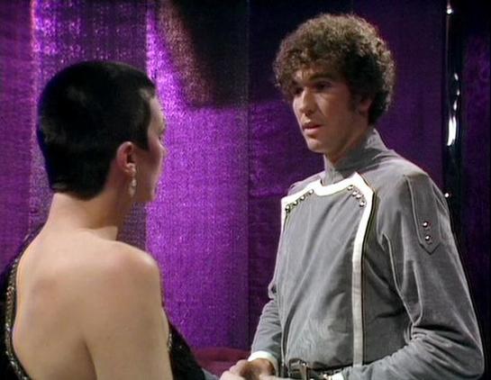 Claude Sonnet 4 describes the image as: "This image appears to be from a scene in the British science fiction television series "Blake's 7," which aired from 1978 to 1981. The setting shows a futuristic interior with distinctive purple/magenta lighting and metallic surfaces, characteristic of the show's space-age aesthetic. Two characters are engaged in what appears to be an intense conversation or confrontation. One figure is shown from behind wearing what looks like a dark, form-fitting outfi…
