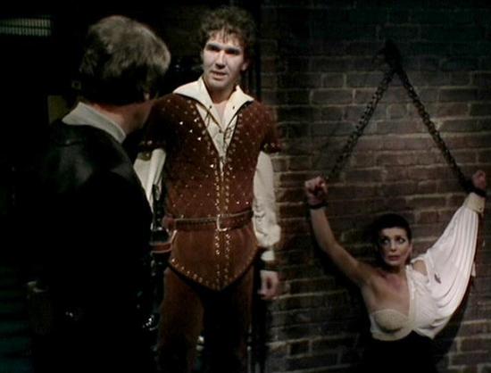 Claude Sonnet 4 describes the image as: "This image appears to be from a dramatic scene in a television production, likely from the science fiction series "Blake's 7" based on the setting and period. The scene takes place in what appears to be a stone dungeon or prison-like setting with brick walls and chains visible in the background.

The setting is dimly lit and atmospheric, creating a tense, dramatic mood. One person is wearing period costume including a brown leather vest or jerkin with de…