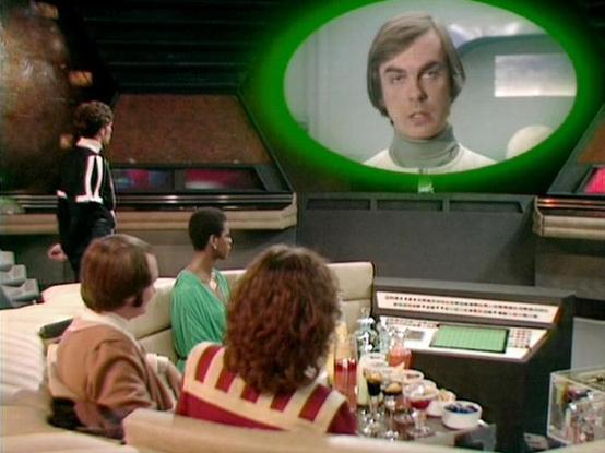 Claude Sonnet 3.7 describes the image as: "This image appears to be from the British science fiction television series "Blake's 7" from the late 1970s/early 1980s. The scene shows a group of people in what looks like a spacecraft control room or bridge. They're seated on light-colored couches/seating areas, watching a large screen or viewscreen with a green glow around it. On the screen appears a person wearing a light turtleneck.

The interior has a distinctive retro-futuristic design typical …