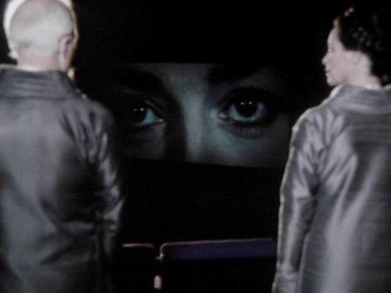 Claude Sonnet 3.7 describes the image as: "The image appears to be a still from a film or television production with a dramatic, noir-like quality. It shows two figures from behind, facing what appears to be a large screen or projection. The two figures are wearing light-colored clothing and stand in silhouette.

In the center of the image, a pair of eyes is visible against a dark background, creating an eerie, watchful presence. The composition creates a sense of tension, with the two figures …