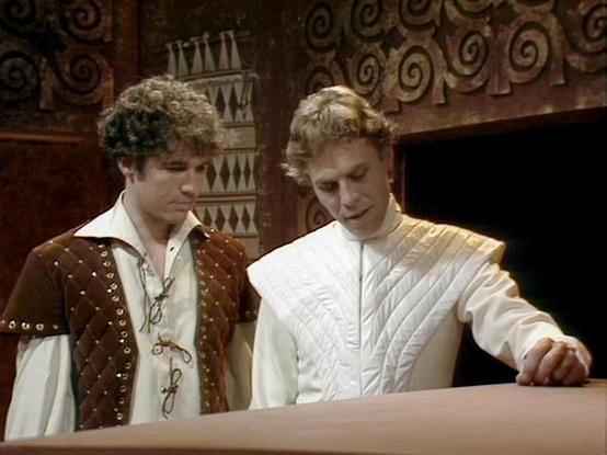 Claude Sonnet 4 describes the image as: "This image shows two actors in period costume, appearing to be from a historical drama or theatrical production. They are dressed in elaborate Renaissance or medieval-style clothing - one wearing a brown leather doublet with decorative studs over a white shirt, and the other in an ornate white quilted or padded garment. The setting appears to be an elegant interior with decorative ironwork visible in the background, suggesting a castle or manor house. Th…